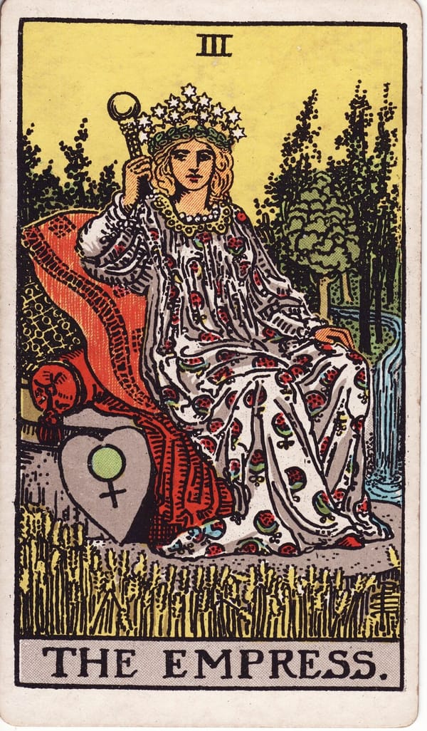 The Empress tarot card: A crowned figure reclines on cushioned luxury in the middle of a ripening wheat field, a river winding through the forest behind her.